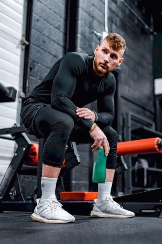 Nam gymer ngồi trên ghế, nhìn xa xăm, tay vòng qua gối, người khom nhẹ khoe cơ tay